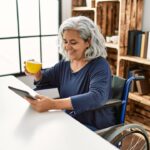 Disabled woman using touchpad and drinking coffee sitting on wheelchair at home.