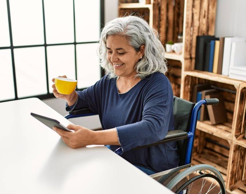 Disabled woman using touchpad and drinking coffee sitting on wheelchair at home.