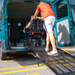 A man guiding a person in a power wheelchair up a metal ramp into the back of a wheelchair-accessible teal van.